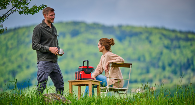 a man and a woman are sitting in a garden with the Einhell cordless coffee maker