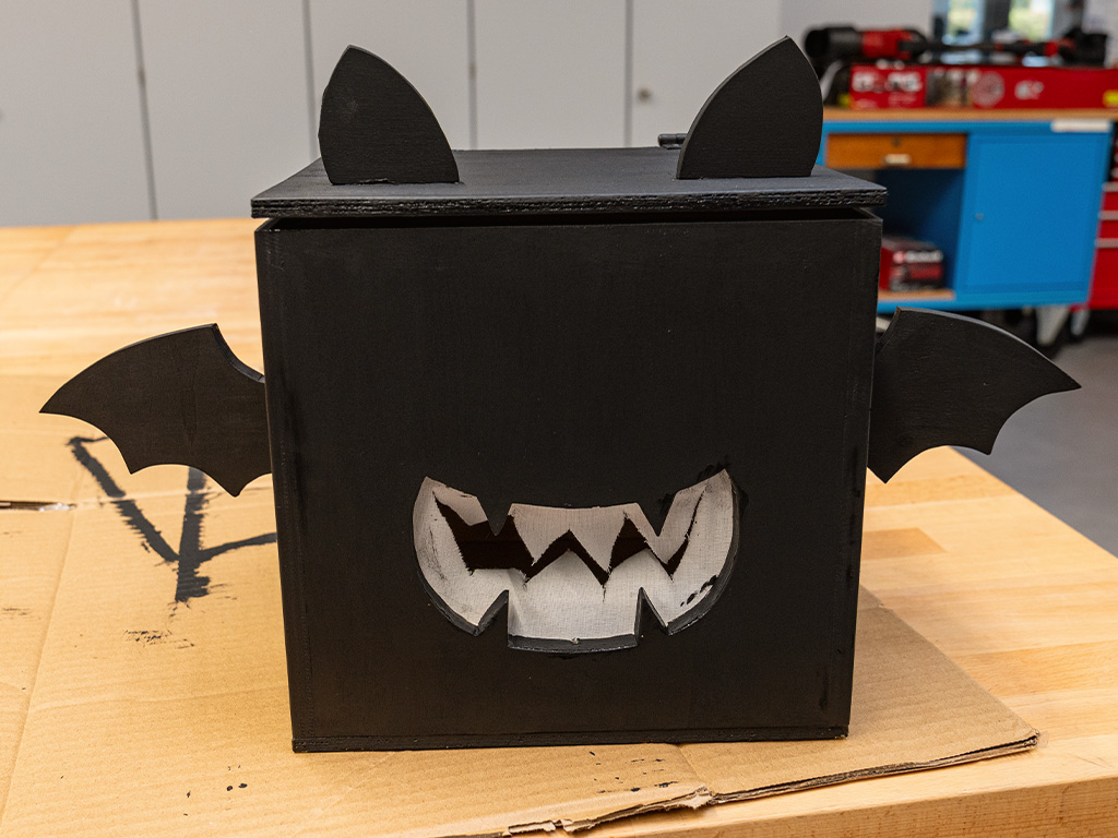 Schwarze Fledermausbox aus Holz mit Ohren, Flügeln und einem weißen, gezackten Maul, die auf einem Stück Pappe auf einer Werkbank steht.