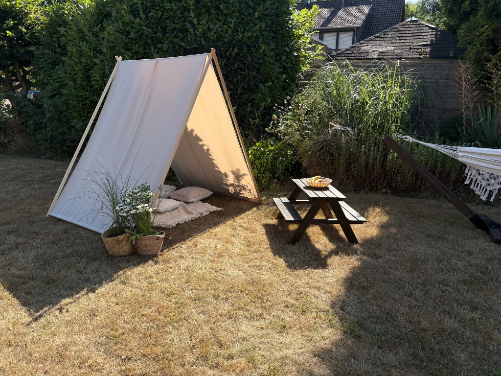 Beige fabric stretched over an A-shaped wooden frame as a sun shelter in a garden, with a picnic table and plants nearby.