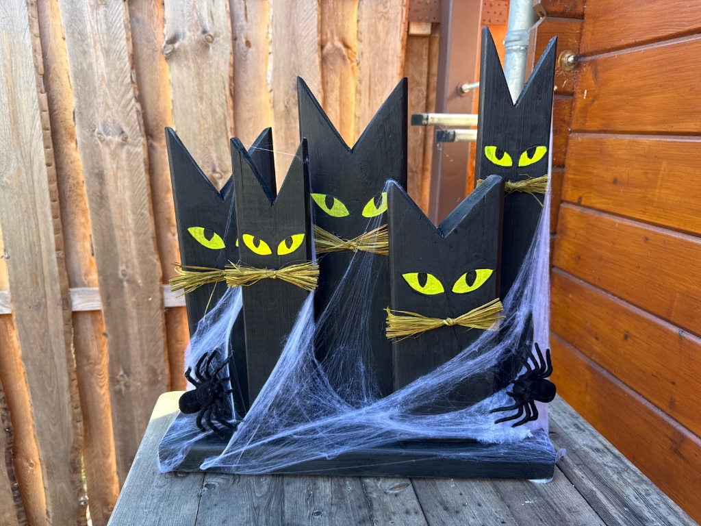 Fünf schwarze, katzenförmige Holzfiguren mit gelben Augen und Schnurrhaaren aus Stroh stehen auf einem gemeinsamen Holzsockel.