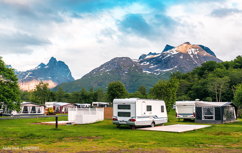 A campsite with caravans and motorhomes against a backdrop of impressive snow-capped mountains and lush green vegetation.