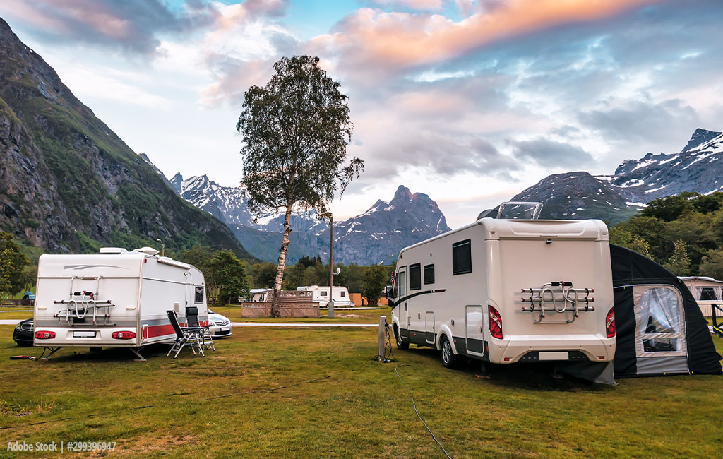 A campsite with caravans and motorhomes in front of a picturesque mountain landscape with snow-covered peaks and a colourful evening sky.