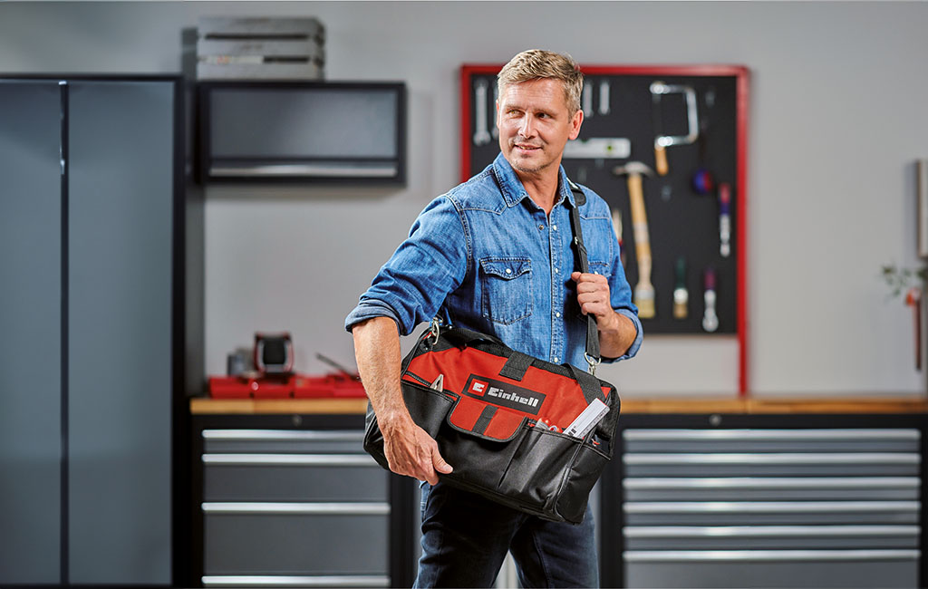 A man in a blue denim shirt carries an Einhell tool bag in a workshop with tools in the background.