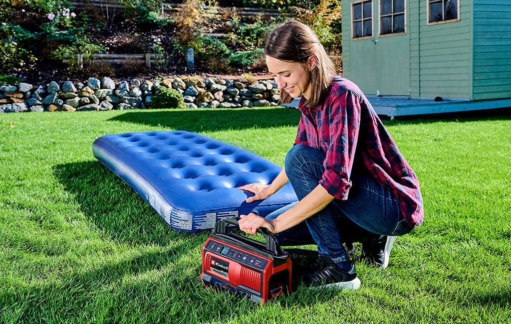 A woman pumps up a blue air mattress with an Einhell air compressor while sitting on a green meadow.