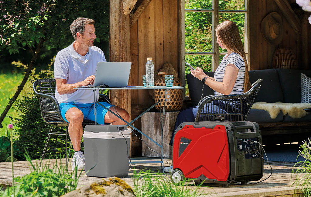 Ein Mann und eine Frau arbeiten auf einer Terrasse im Freien, beide Geräte sind an einen Einhell-Generator angeschlossen, der auf dem Boden neben ihnen steht.