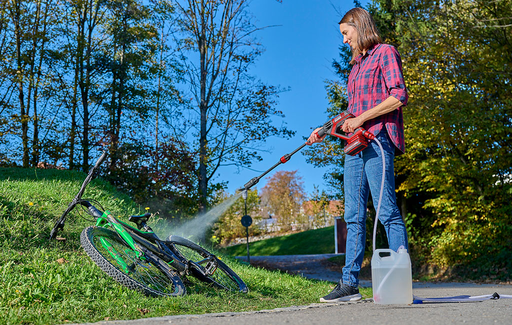 A woman cleans a green bicycle with an Einhell high-pressure cleaner on a grassy hill on a sunny day.