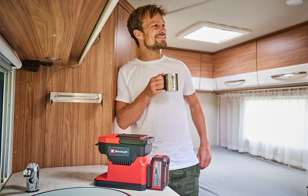 A man stands in a mobile home, holding a cup and smiling, while the Einhell cordless coffee maker is on the kitchen counter in front of him.