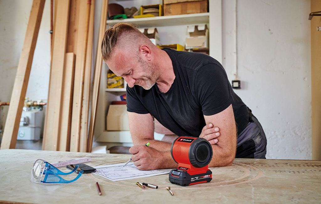 A man is working in a workshop, leaning over a table and writing on a sheet of paper, while an Einhell speaker and safety goggles lie next to him.