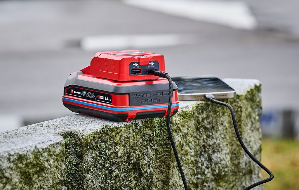 An Einhell SEALED battery charges a smartphone via a USB cable while it is lying on a concrete block outdoors.