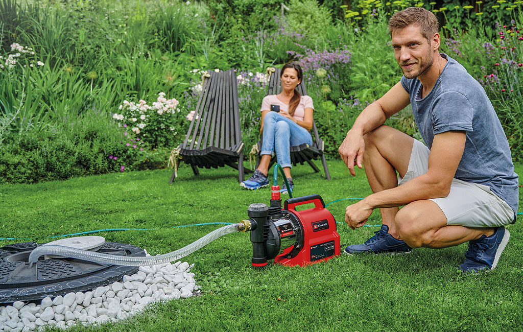 Ein Mann kniet neben einer Gartenpumpe, während eine Frau im Hintergrund sitzt.