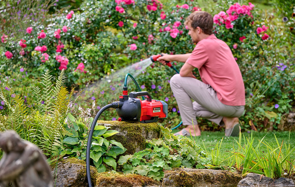 A man kneels in the garden, using a Einhell garden pump to water plants.