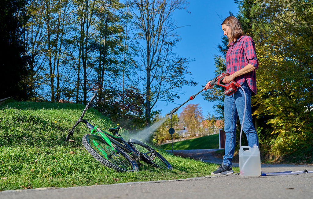 Eine Frau in kariertem Hemd und Jeans reinigt ein Fahrrad mit einem Hochdruckreiniger im Freien bei sonnigem Wetter.