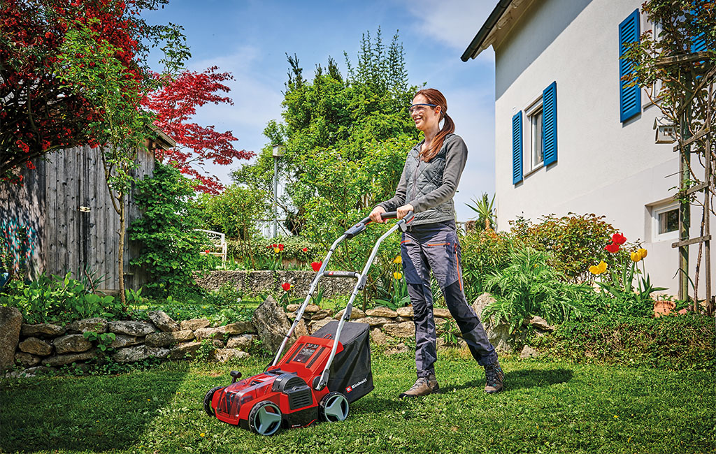 Eine Frau beim Rasenlüften mit einem Akku-Vertikutierer-Lüfter im Garten vor einem Haus.