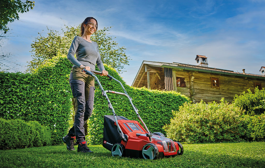 Eine Frau beim Rasenlüften mit einem kabellose Vertikutierer-Lüfter im Garten vor einem Holzhaus.