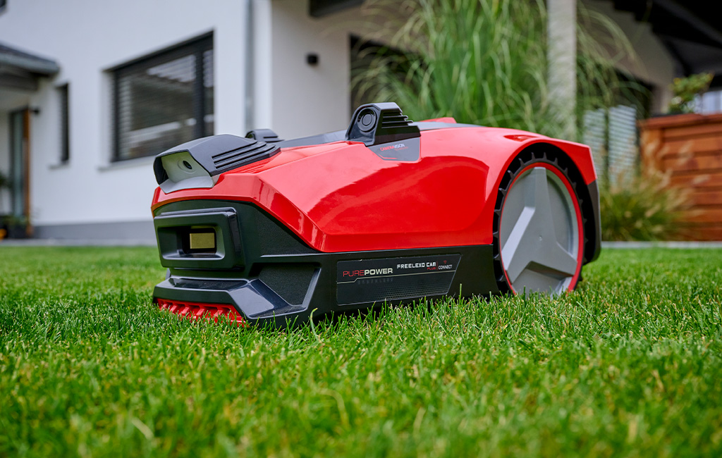 A red Einhell robotic lawnmower stands on a well-maintained lawn in front of a modern house.