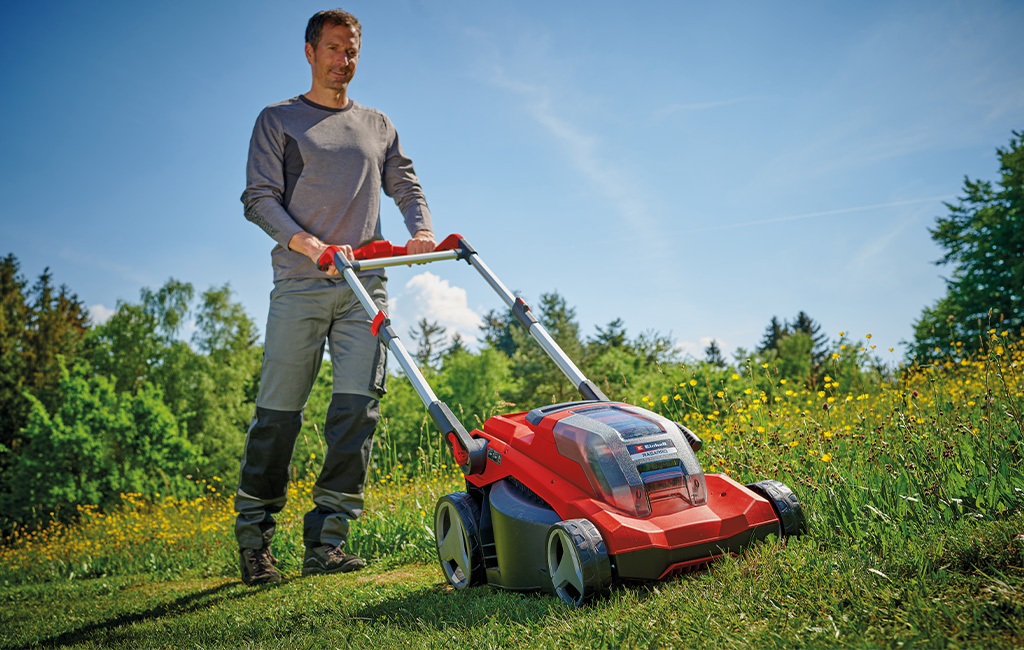 A man is mowing a blooming meadow with a red Einhell lawnmower under a sunny, blue sky.