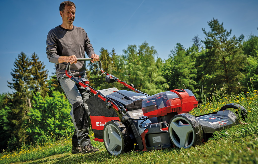 A man is mowing a green meadow on a slope with an Einhell cordless lawnmower.