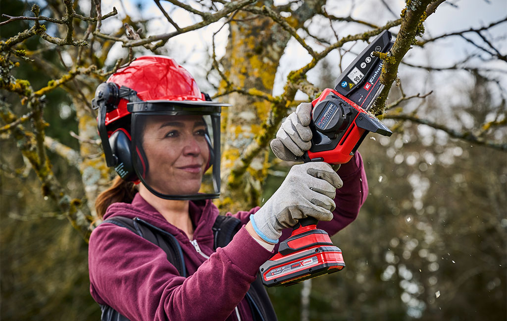 Frau mit Forstschutzhelm sägt den Ast eines Baumes mit einer Akku-Astkettensäge ab.