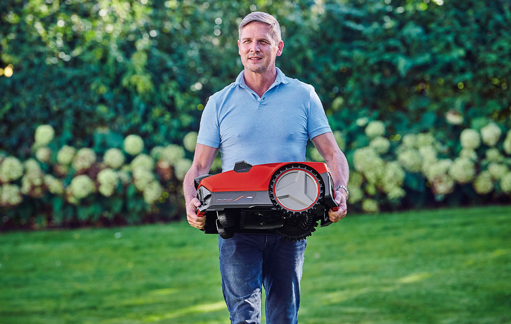 A man carries a robotic lawnmower through the garden.