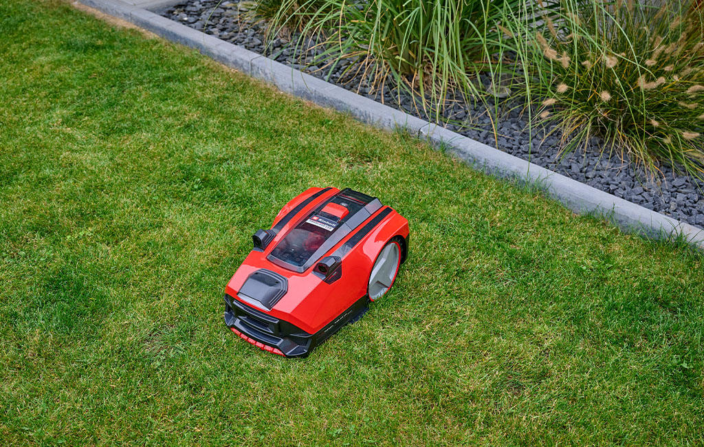 An Einhell FREELEXO CAM robotic lawnmower on a green lawn in front of a garden bed