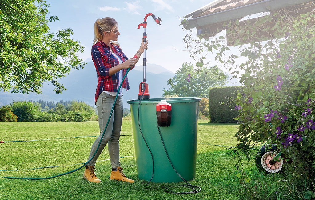 Eine Frau benutzt eine Einhell Akku-Tauchpumpe, um Wasser aus einer Regentonne in einem sonnigen Garten mit Blick auf die Berge zu pumpen.