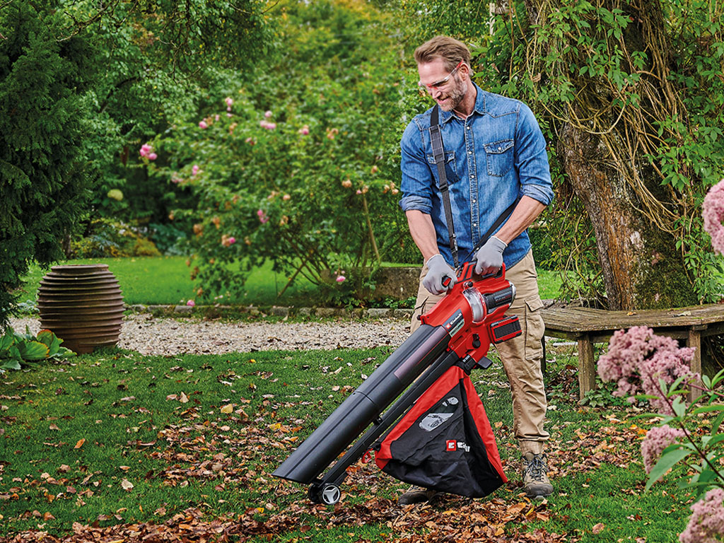 A man uses a Einhell leaf vaccum to clear leaves in an autumn garden, surrounded by blooming plants and trees.