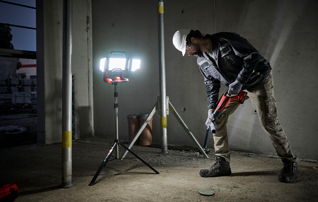 A construction worker uses an Einhell cordless work light on a tripod to illuminate a dark construction site