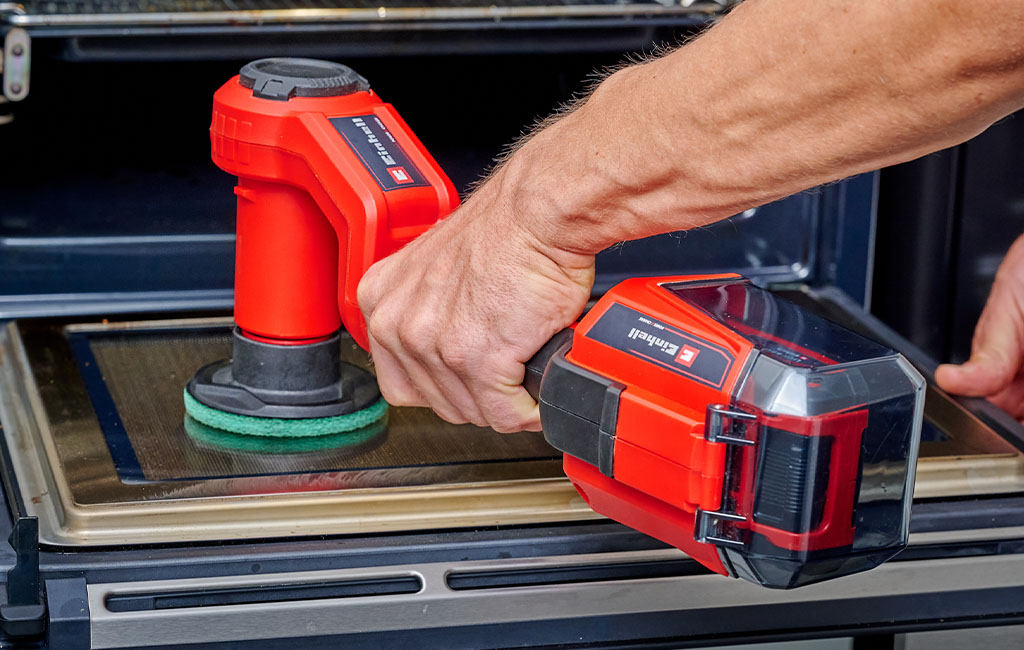 An Einhell cordless surface brush being used to clean a stove glass panel.