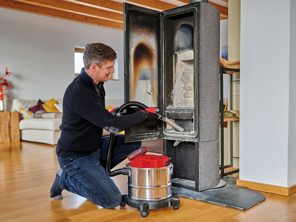 A man cleaning a wood-burning stove with an ash vacuum in a modern living room.