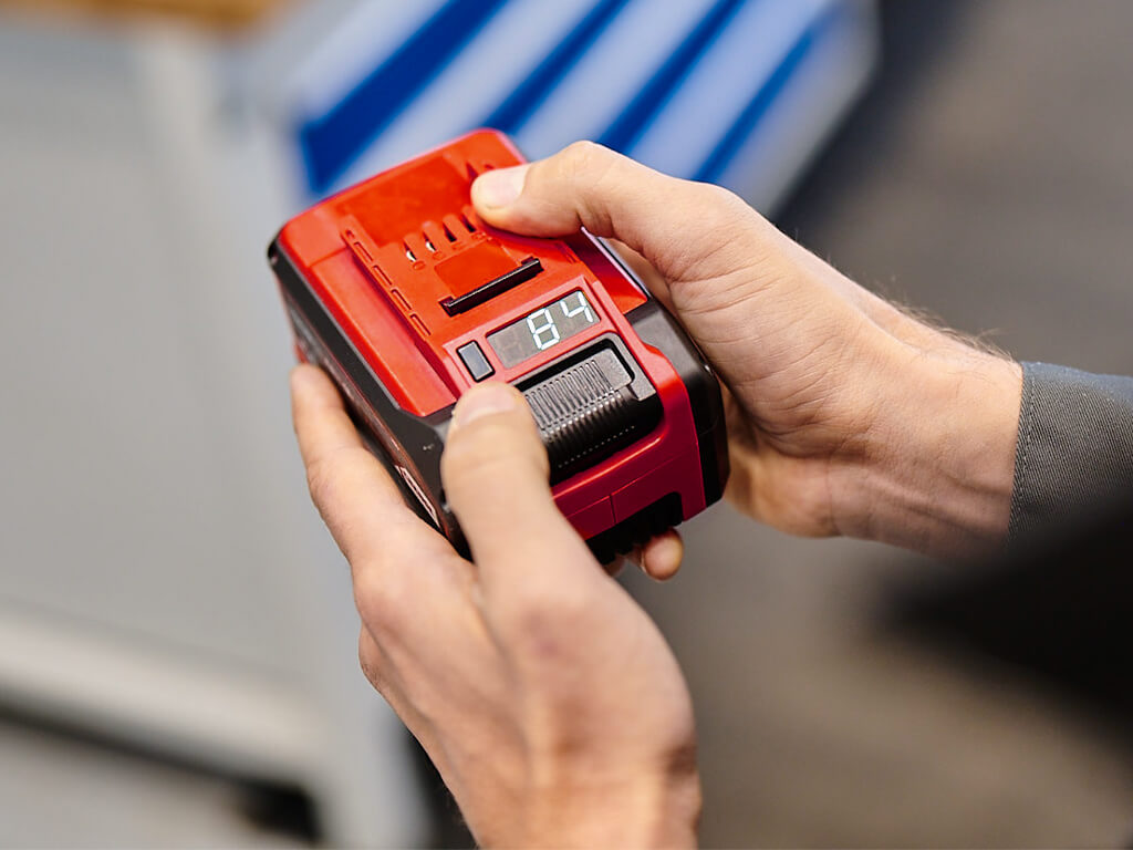 Two hands hold a Power X-Change battery, on which the LED display shows 84% charge status.