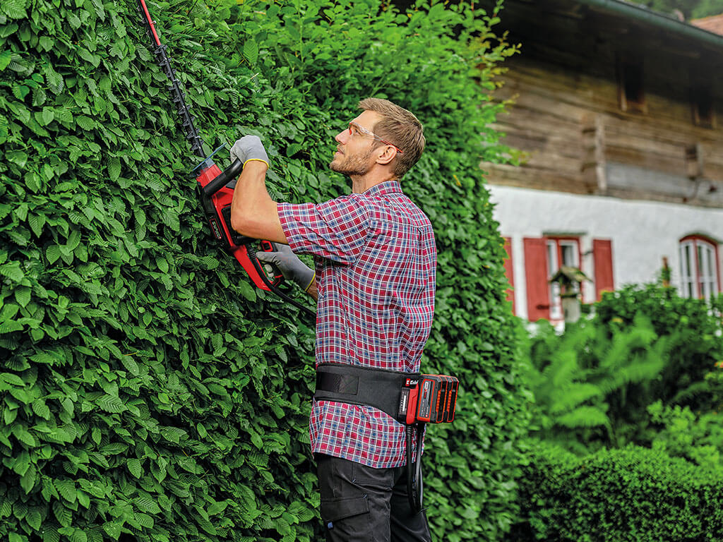 A man working with a cordless hedge trimmer while wearing the two Power X-Change batteries in the battery belt on his hip.