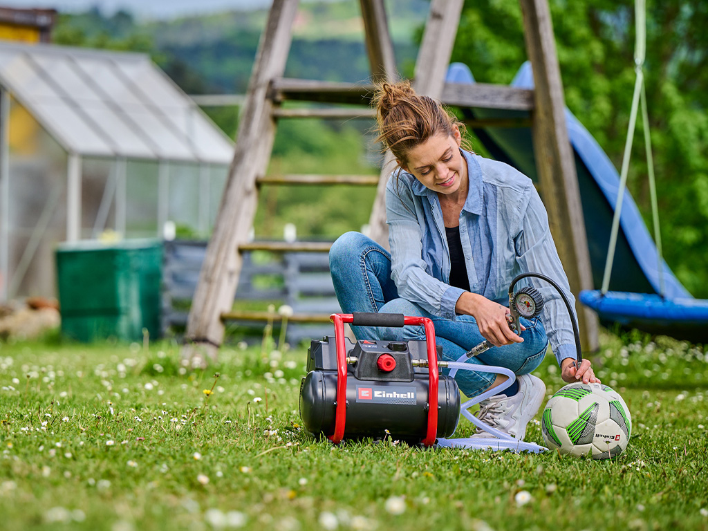 Eine Frau benutzt einen tragbaren Einhell-Kompressor im Garten, um einen Fußball aufzupumpen.