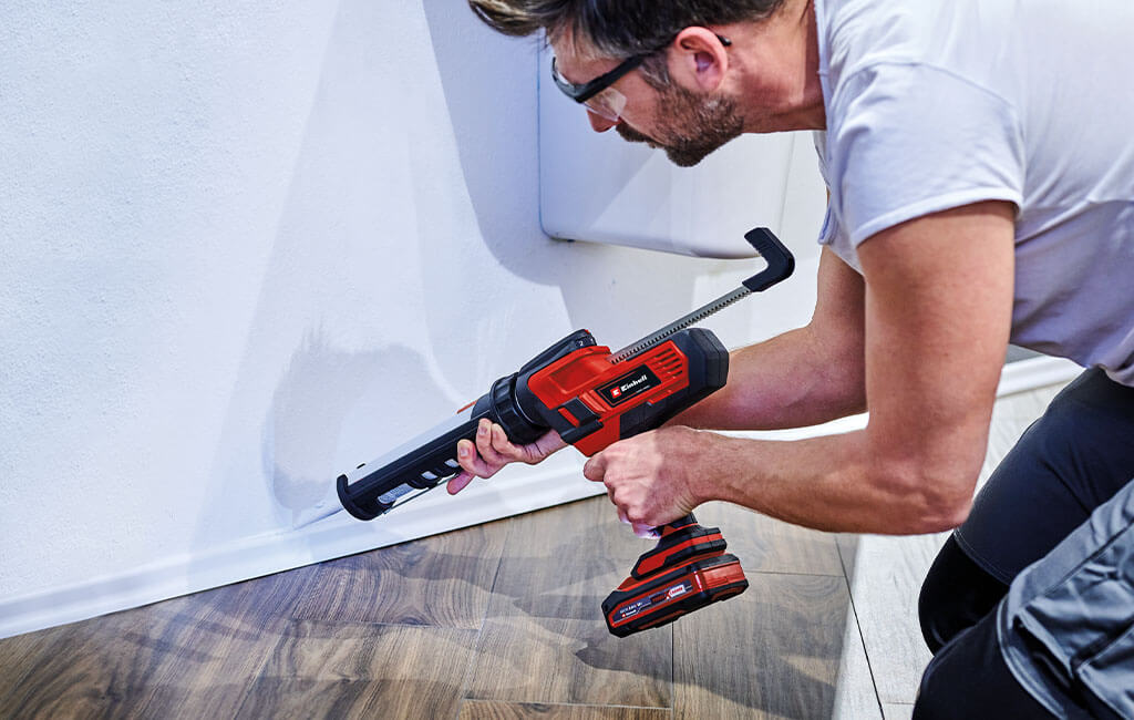 A man kneeling on the floor is pulling a silicone sealant along the baseboard.