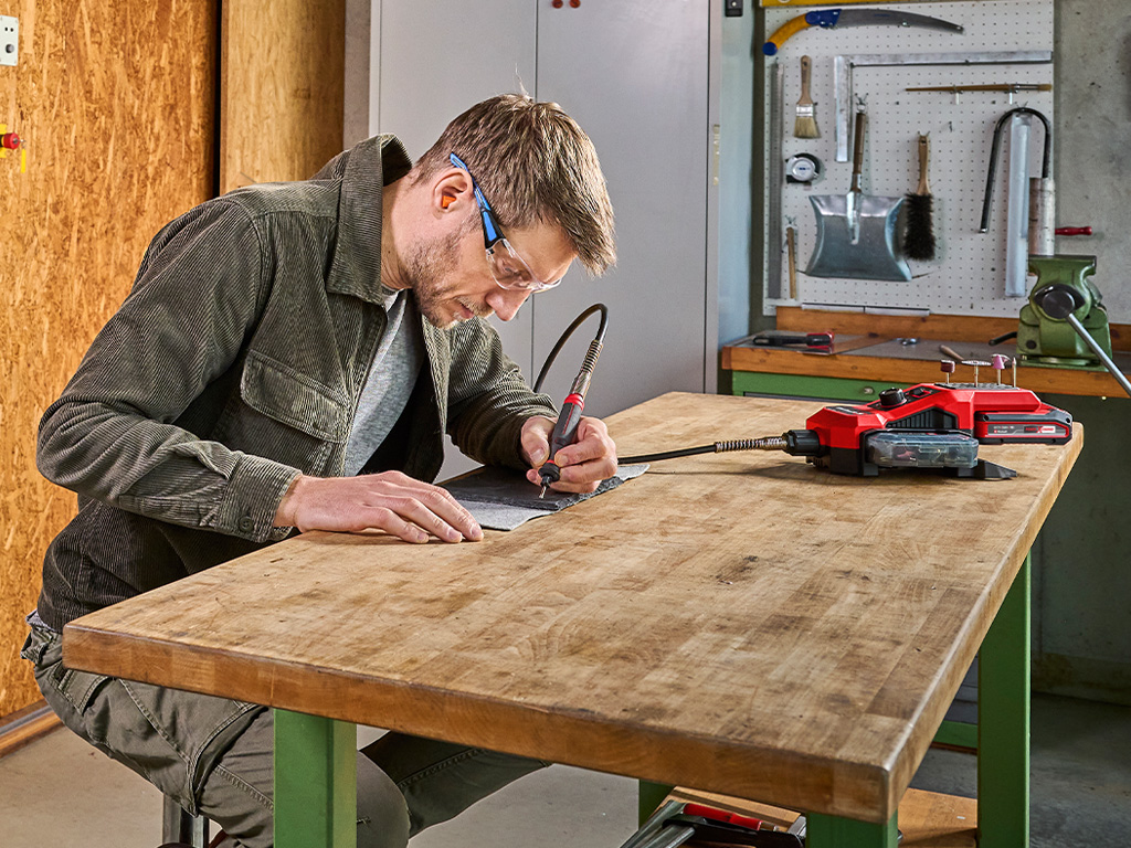 A man engraves a metal plate on a workbench using a red, flexible engraving tool from Einhell.