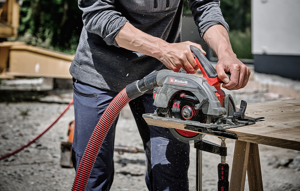 A person using a handheld circular saw from Einhell connected to a vacuum is cutting wood.