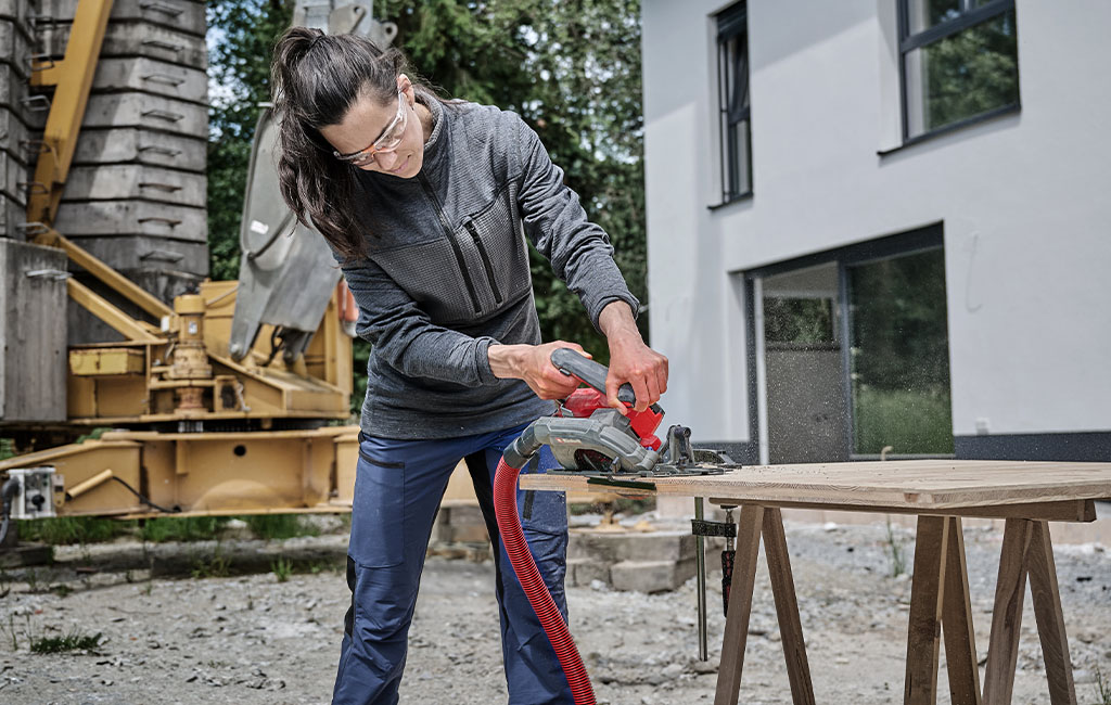 A woman wearing safety glasses uses a handheld circular saw to cut wood at a construction site.