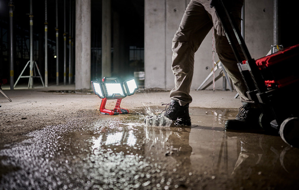 A portable LED work light stands in water at a construction site as a person walks by.