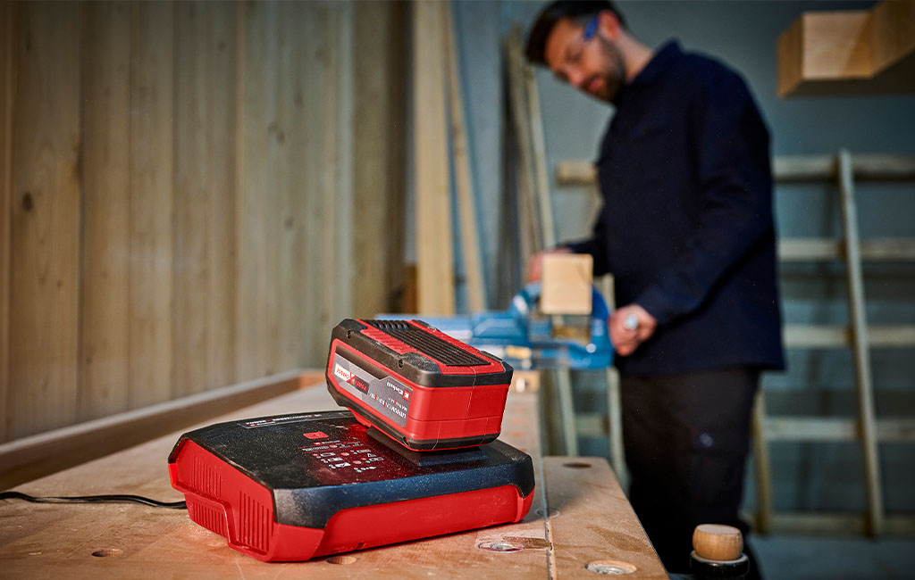 im Vordergrund ein Einhell Boost-Charger mit Power X-Change Akku; im Hintergrund ein Mann, der mit Holz arbeitet