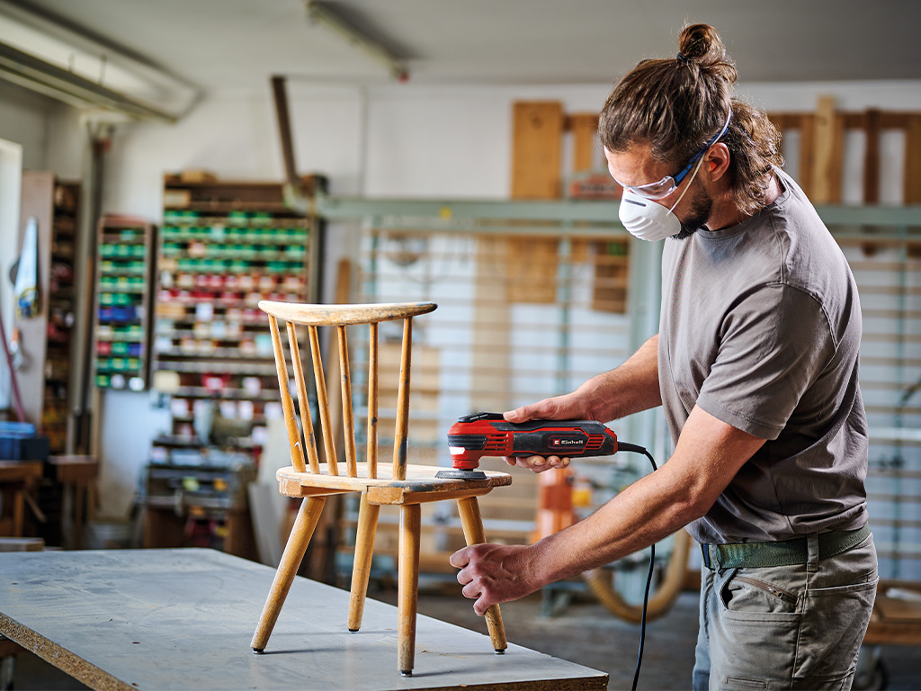 A man sands a small, old wooden chair with a multitool in a workshop, wearing safety goggles and a mask.